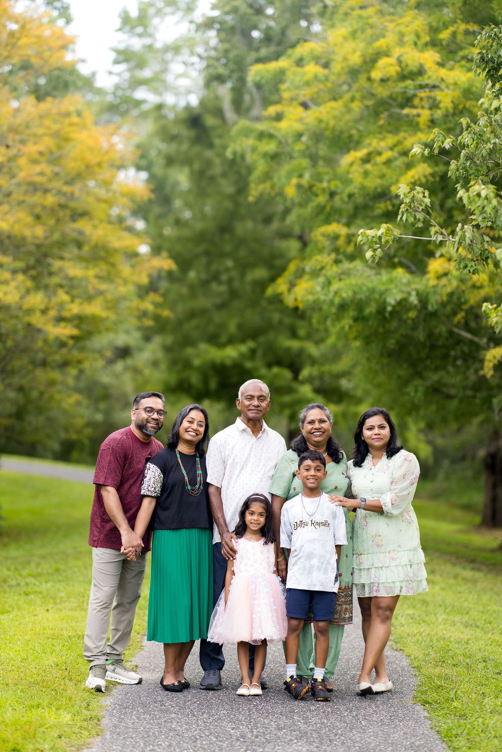 Multi-generational South Asian family posing together on a park path surrounded by lush green trees.