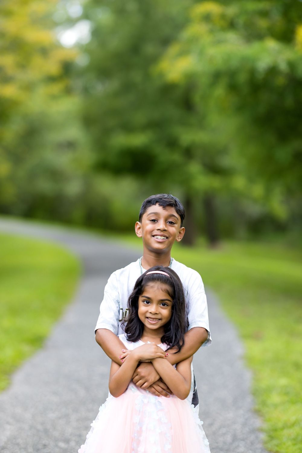 Two Indian siblings pose on a park path, older boy hugging younger girl in pink tutu dress, surrounded by lush greenery.