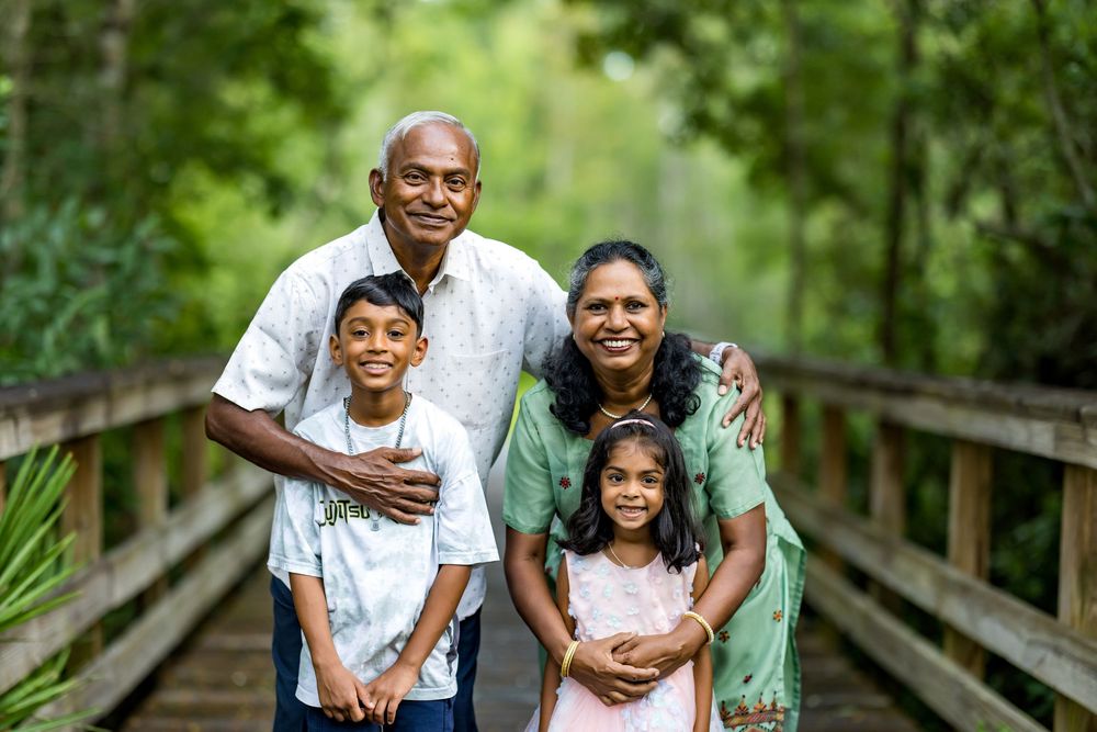 Multi-generational South Asian family of four smiling together on a wooden bridge surrounded by lush green trees.