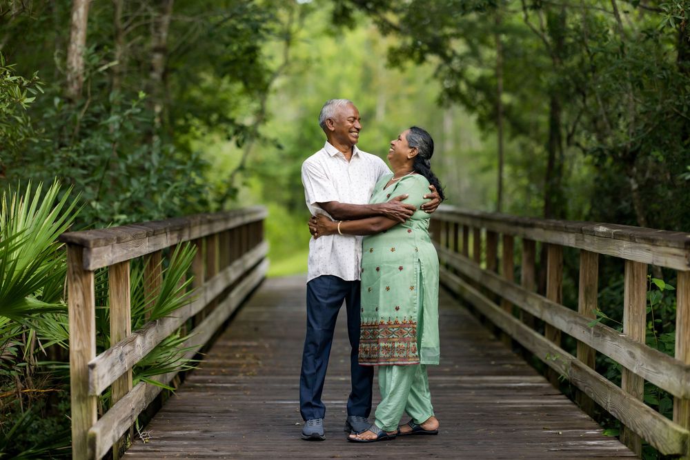Elderly Indian couple embracing and sharing a romantic moment on a wooden bridge surrounded by lush green trees.