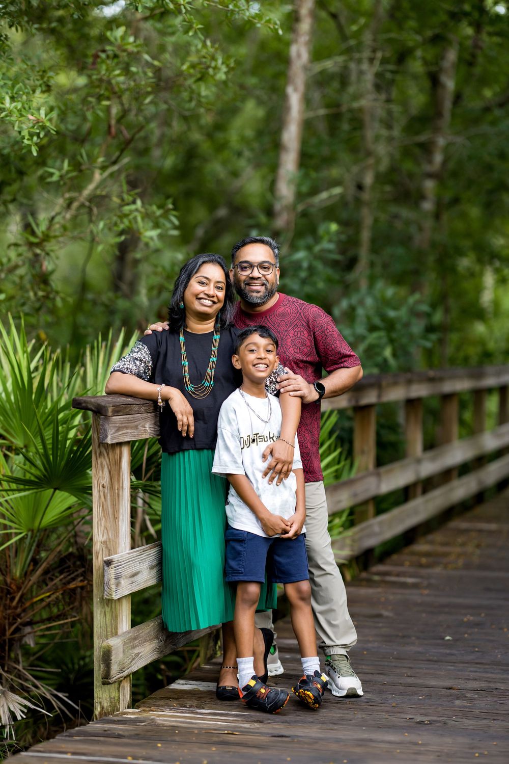 A smiling family of three poses on a wooden bridge surrounded by lush green trees and tropical plants.