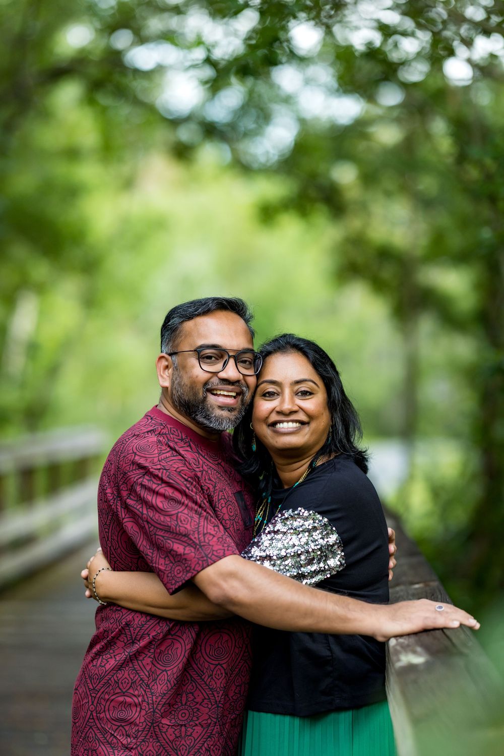 A smiling couple embraces on a wooden bridge surrounded by lush green trees in an outdoor park setting.