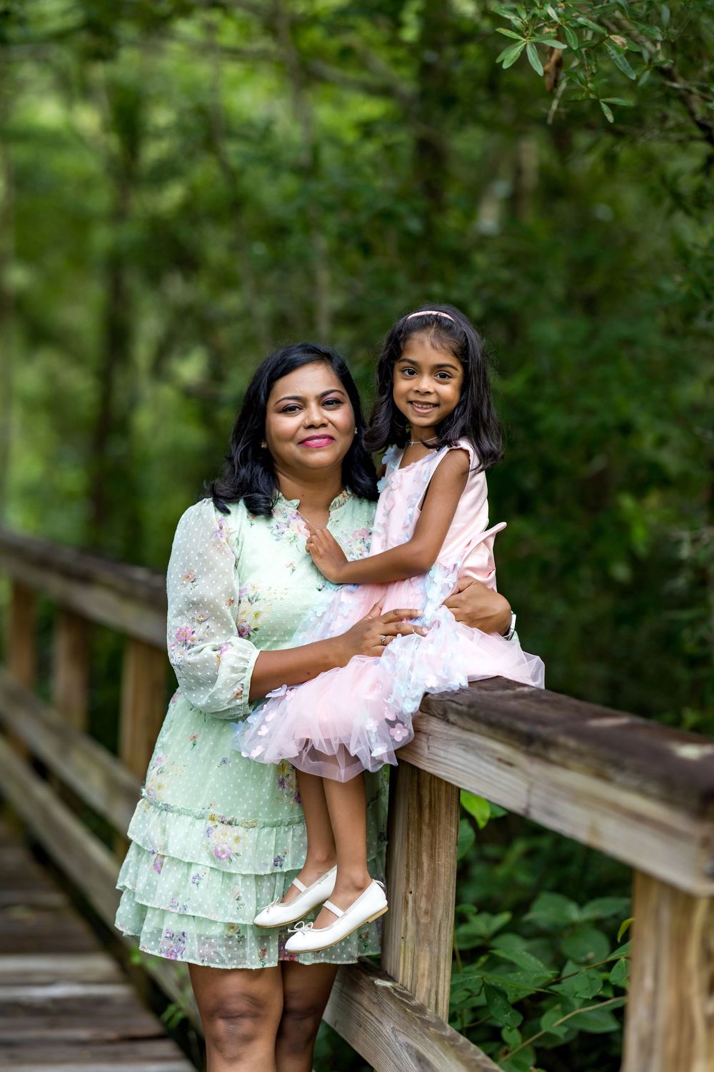 A mother and young daughter in matching pink and green dresses pose on a wooden bridge surrounded by lush greenery.