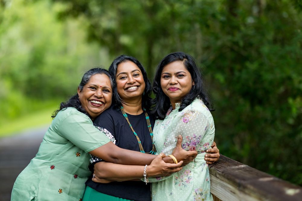 Three happy South Asian women hugging and smiling outdoors on a green tree-lined path.