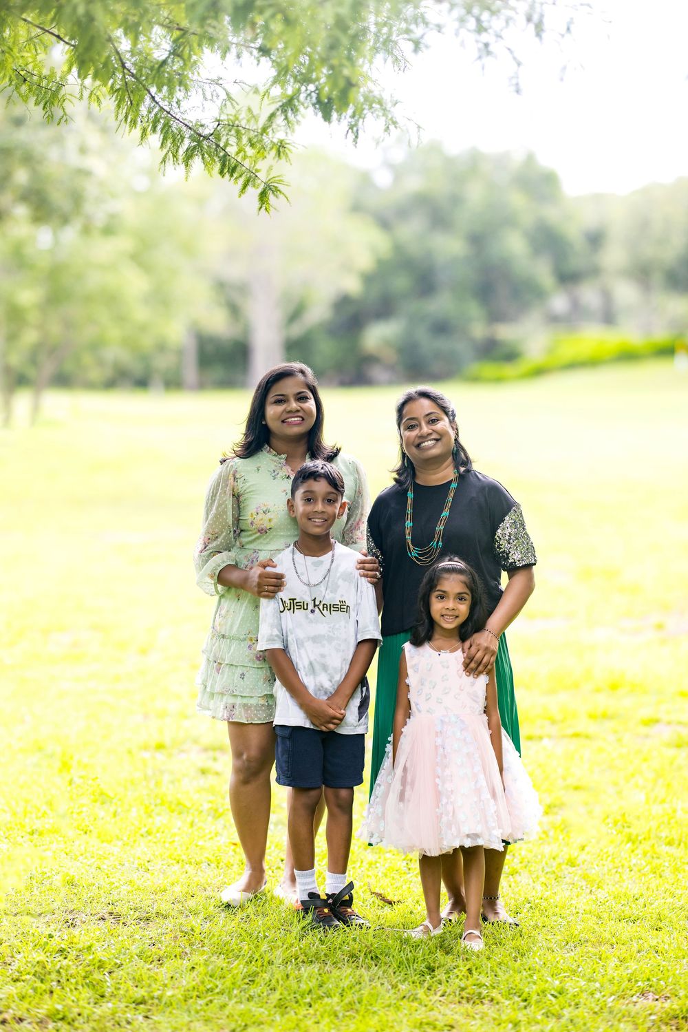 A joyful South Asian family of four posing together on green grass in a sunny park setting.