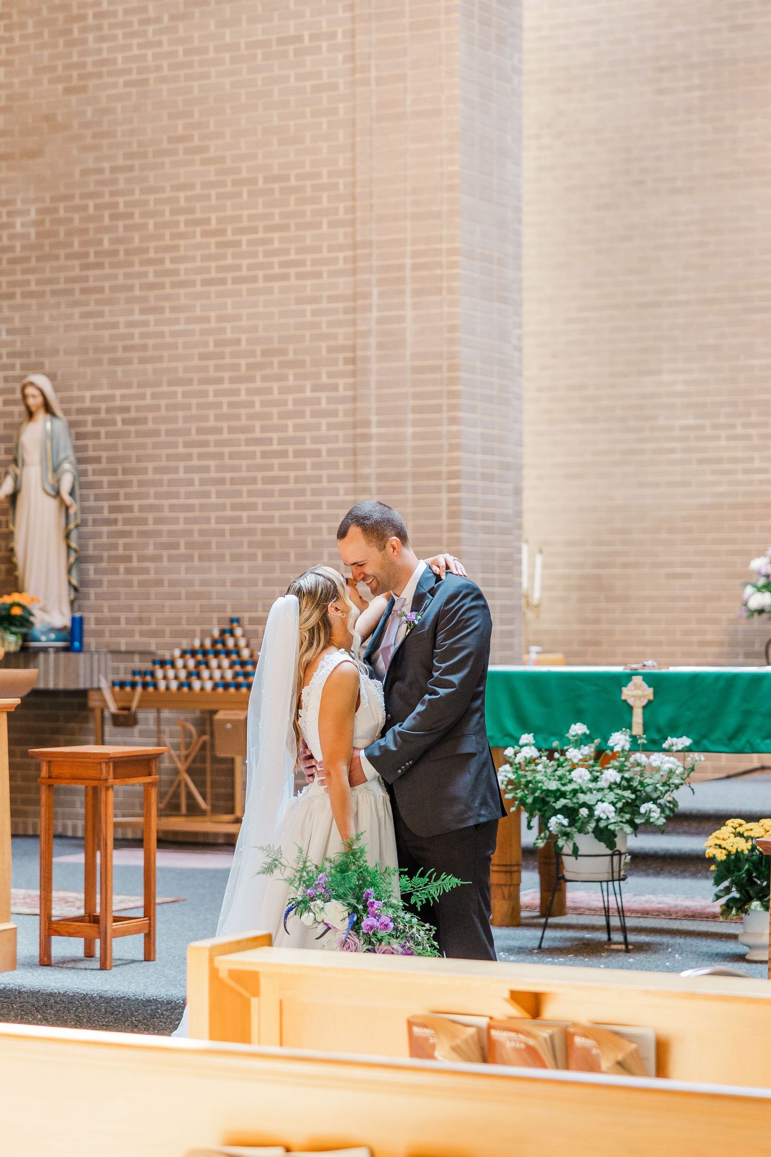 Bride and groom share a romantic first kiss at the altar of a brick church, surrounded by floral arrangements and wooden pews.