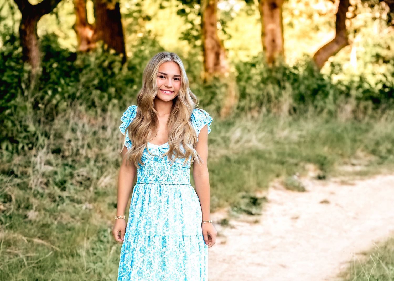 Young woman in blue floral maxi dress smiling in golden field at sunset.