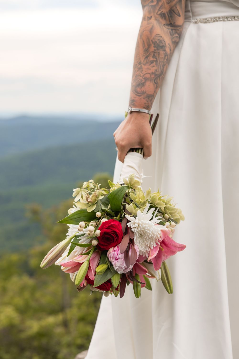 Tattooed bride in white dress holding a colorful wedding bouquet with roses and lilies, mountain landscape in background.
