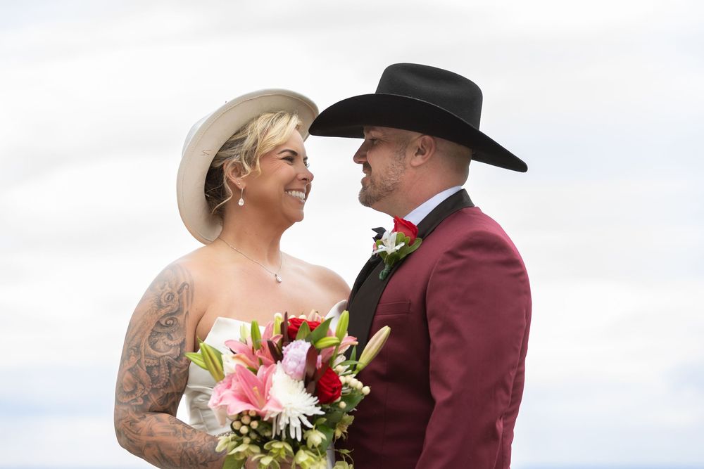 Bride with tattoos in white dress and groom in burgundy suit with black cowboy hats exchange loving glances at outdoor wedding.