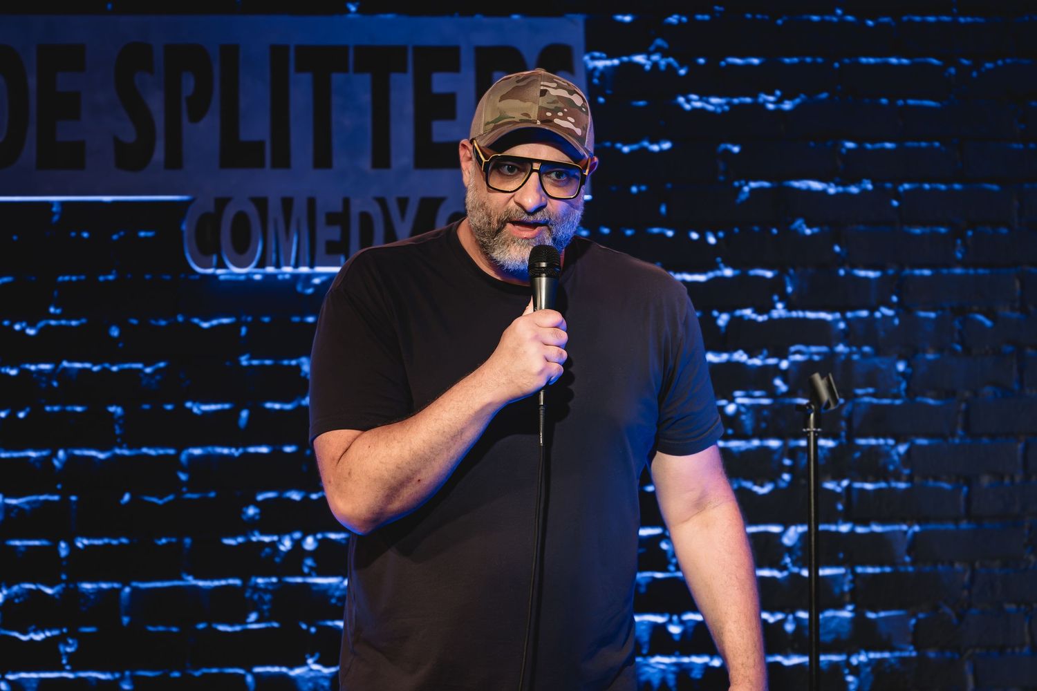 Kurt Metzger, a stand-up comedian performs on stage at Side Splitters Comedy Club with a brick wall behind him.