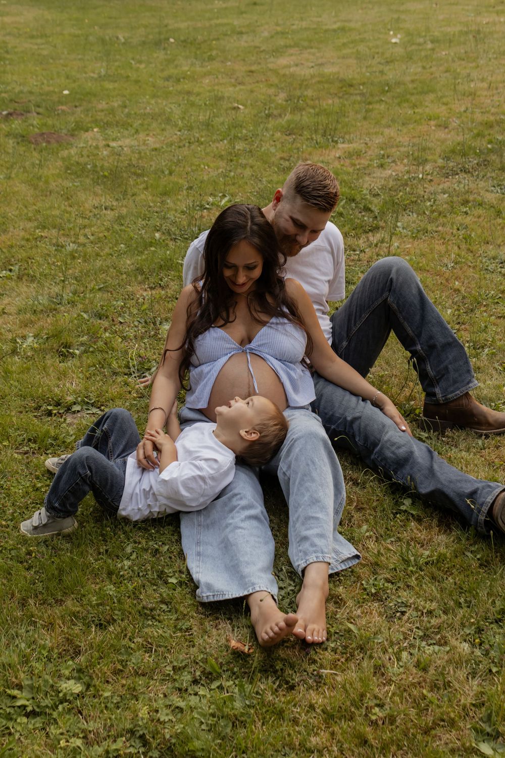 Pregnant woman sits between partner and child on grass, family maternity photo outdoors.