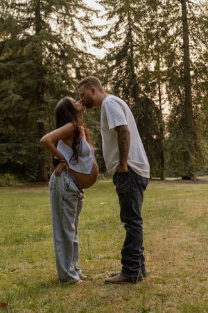 Couple sharing a kiss outdoors in a park, with pregnant woman showing baby bump, surrounded by tall evergreen trees.