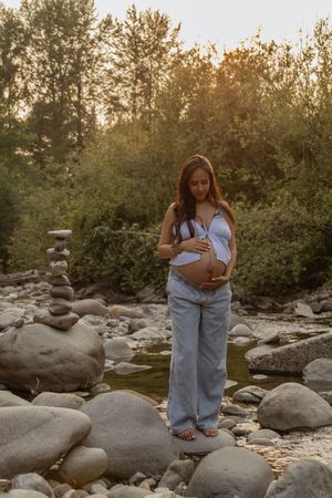 Pregnant woman cradling her belly outdoors on rocky riverbank at golden hour with lush green trees on little mashel river