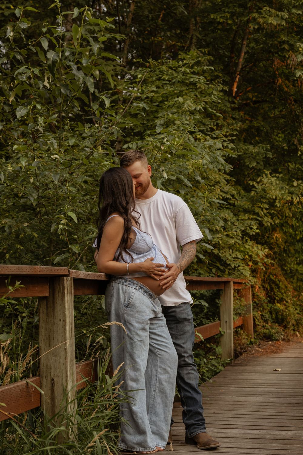 Couple shares tender kiss on wooden bridge, man holds pregnant womans belly in eatonville WA.