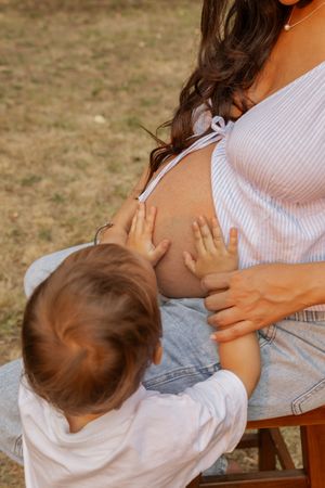 A toddler touching a pregnant woman's bare belly outdoors in a sunny park setting.