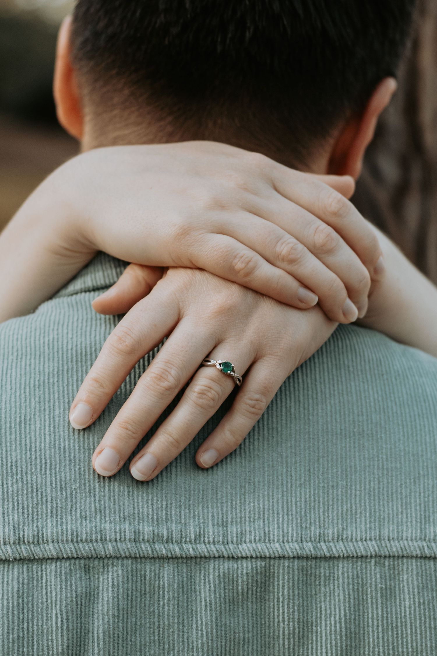 Close-up of a womans hand with a silver and emerald engagement ring wrapped around her partners shoulder.