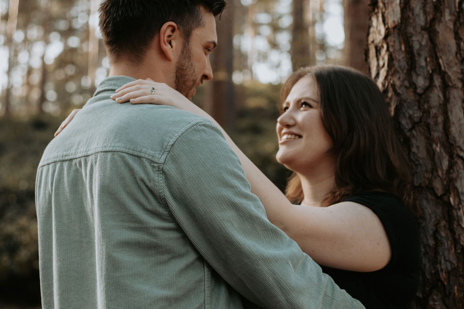 A couple embraces romantically among tall pine trees, the woman smiling warmly at her partner outdoors.