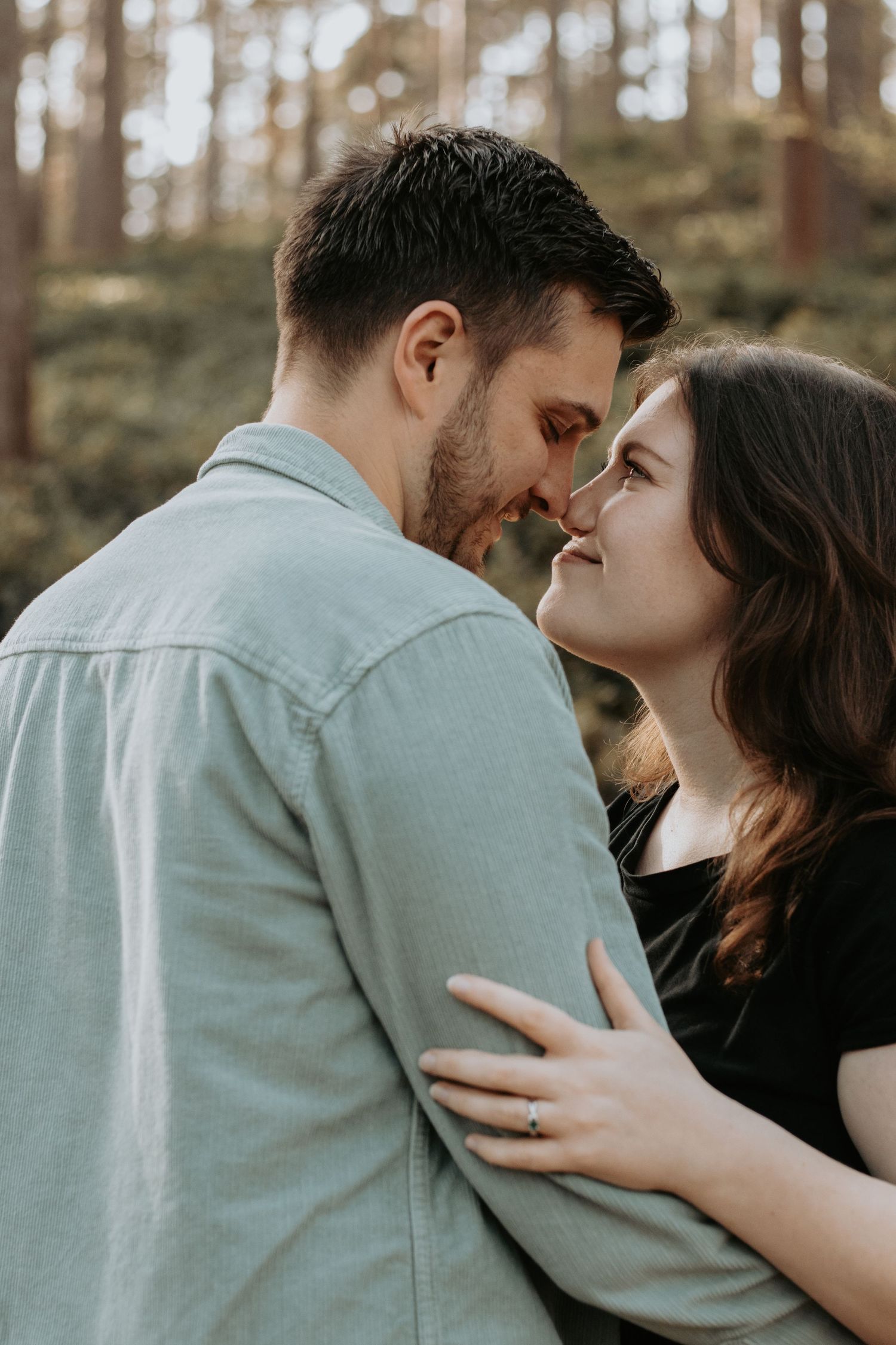 Couple sharing a romantic moment in a forest, woman wearing engagement ring embracing man in light blue shirt.