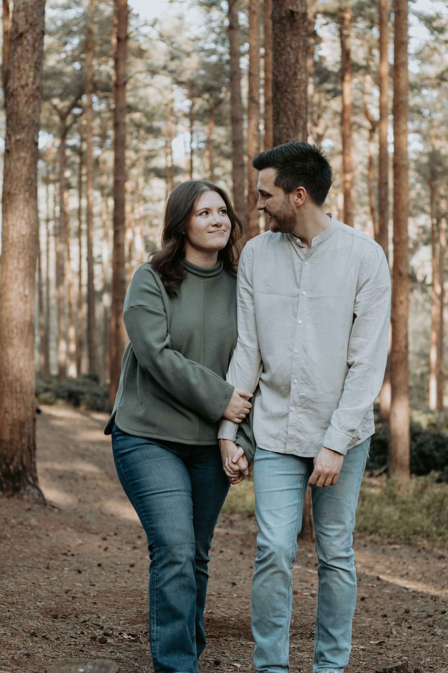 A couple walks hand-in-hand through a pine forest, smiling at each other, wearing casual jeans and tops.