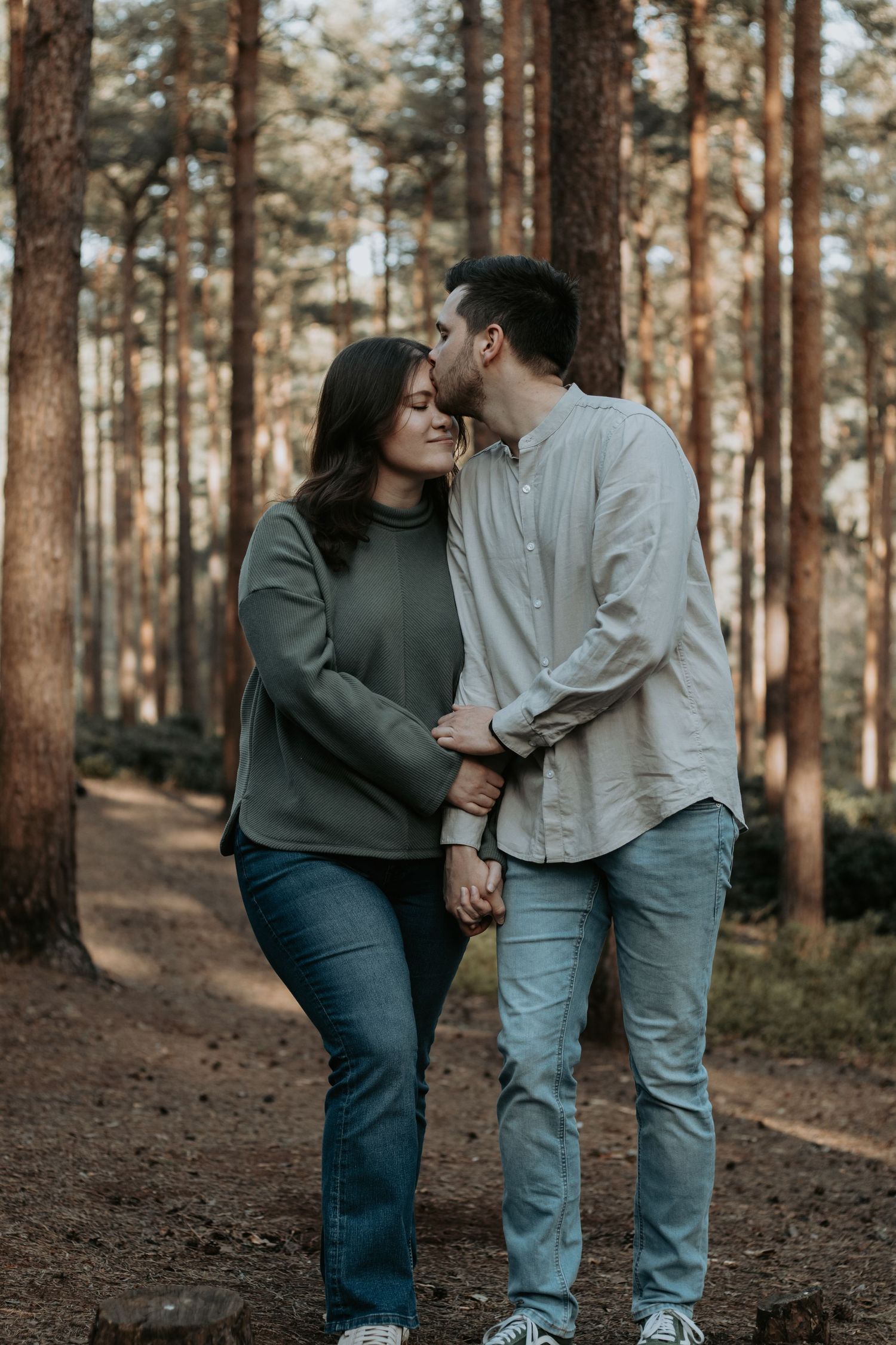 Couple sharing a romantic kiss in a pine forest, holding hands while the woman smiles in a green sweater and jeans.