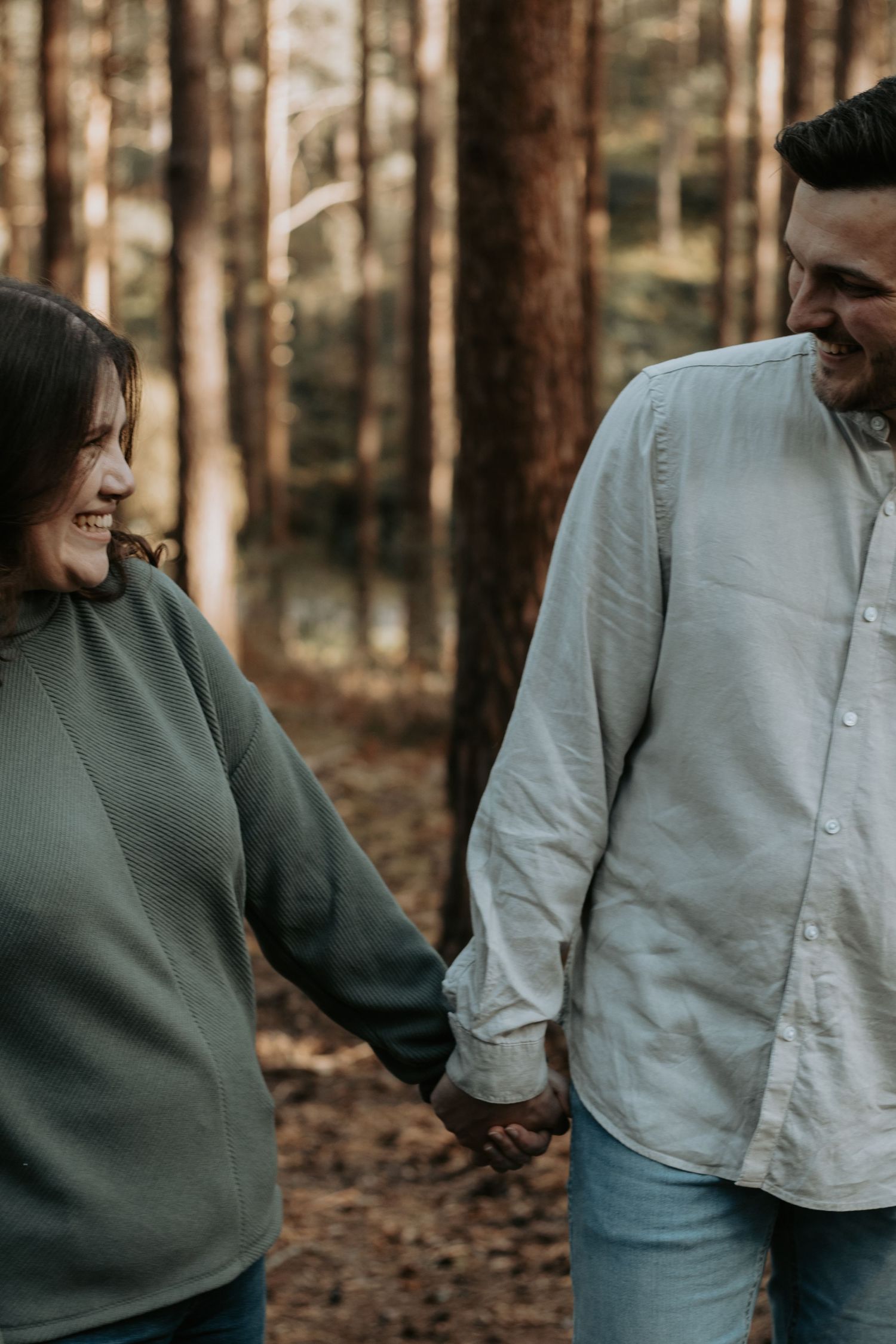 A smiling couple holds hands while walking through a sunlit pine forest, dressed in casual green and white tops.