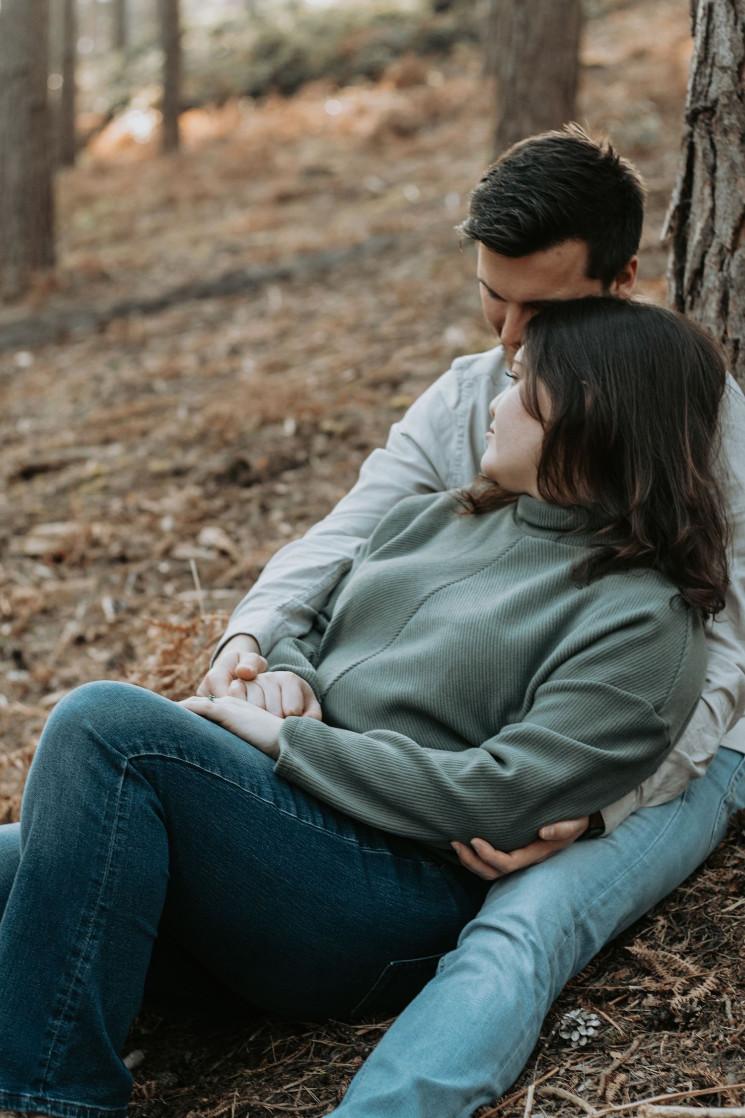 Couple sitting against a tree in a forest, sharing a tender moment while embracing each other warmly.