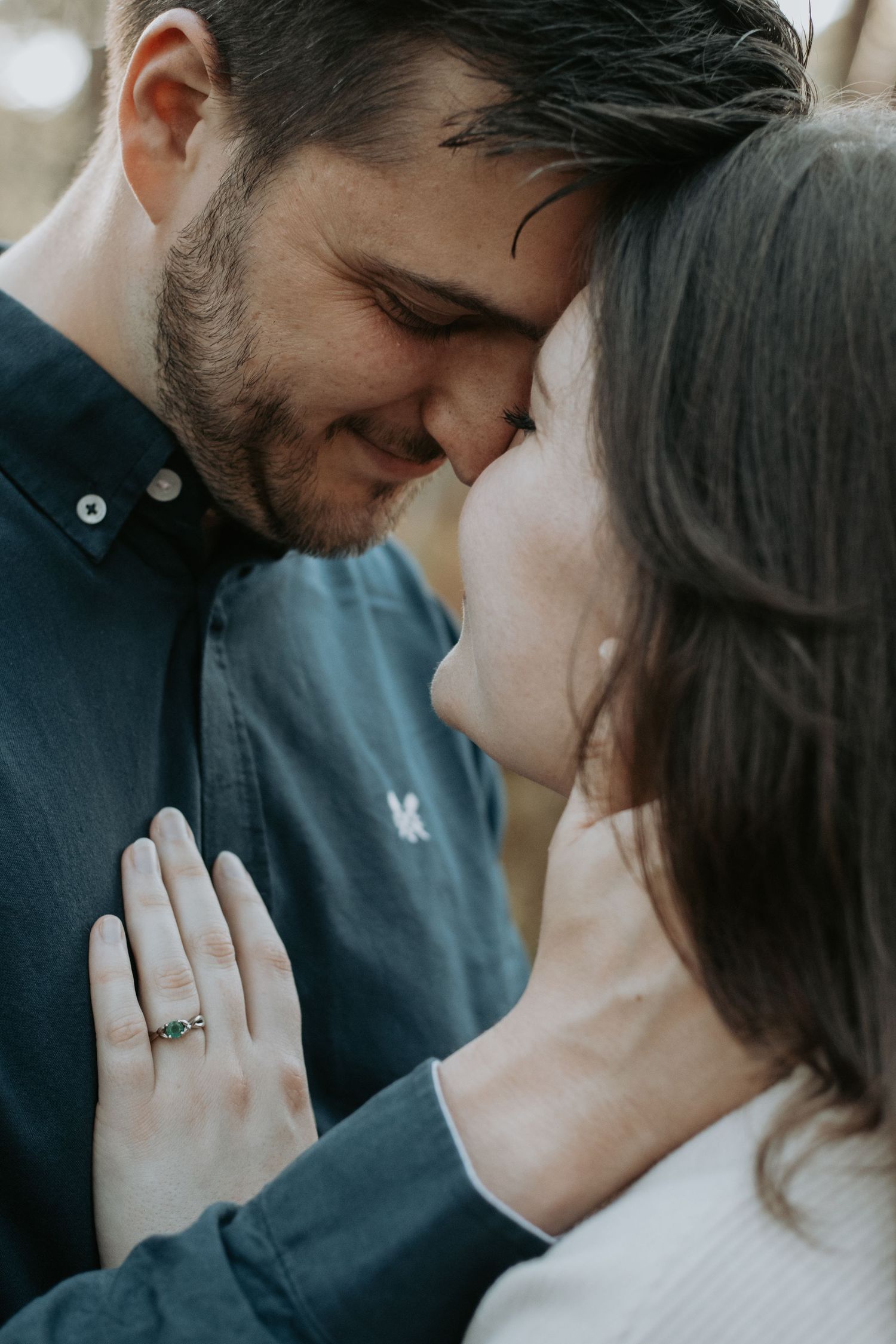Couple sharing a romantic forehead touch, woman wearing an emerald engagement ring, soft natural light outdoor setting.