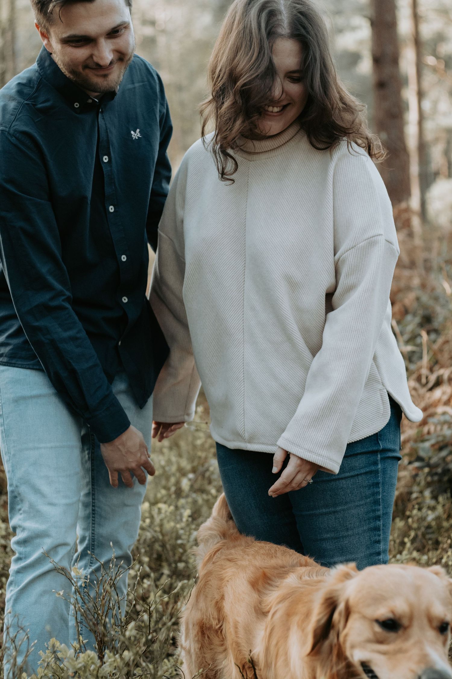 A smiling couple walks with a golden retriever through a forest, woman in cream sweater and man in dark shirt.