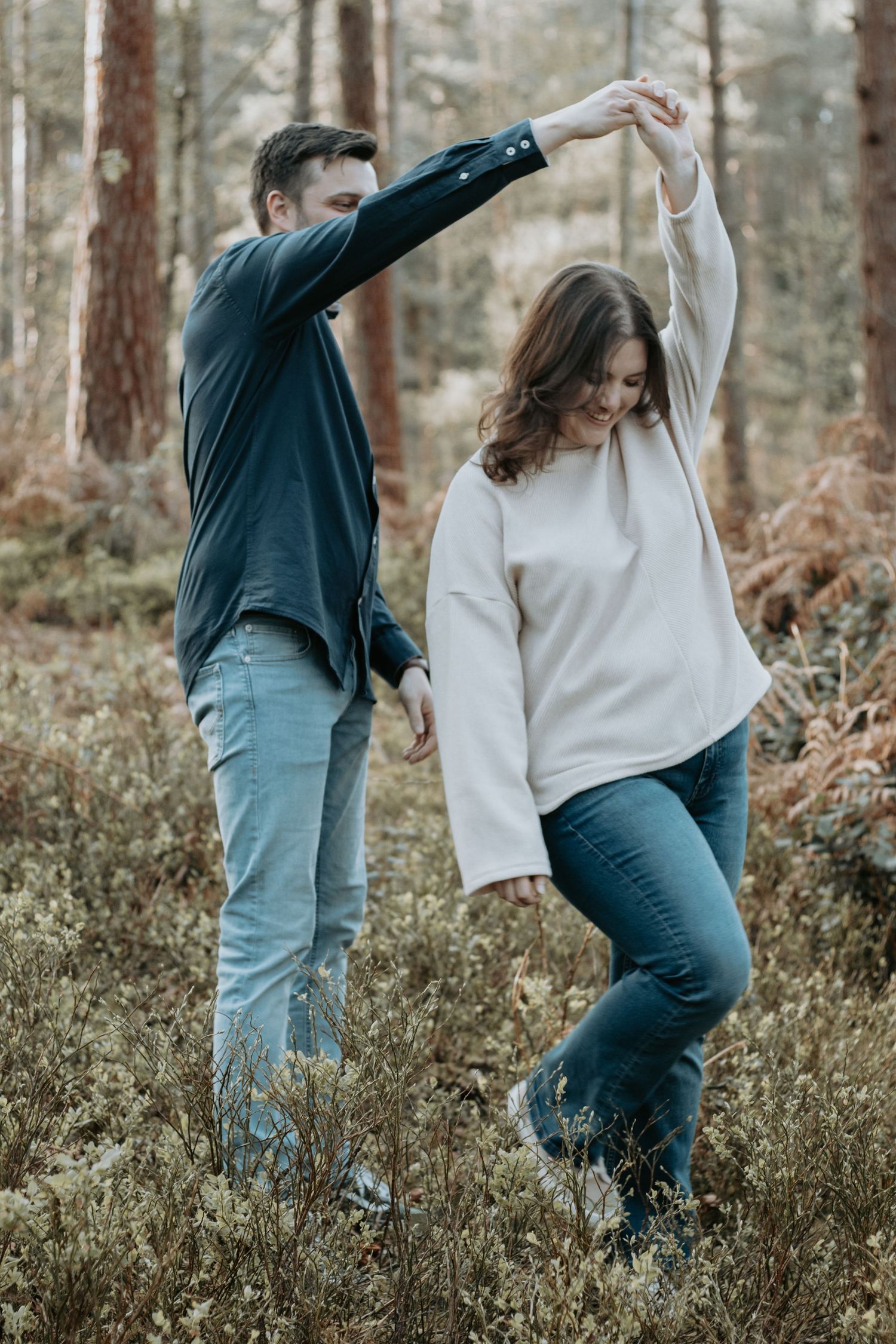 Couple dancing playfully in a forest, man spinning woman gently among autumn foliage and pine trees.