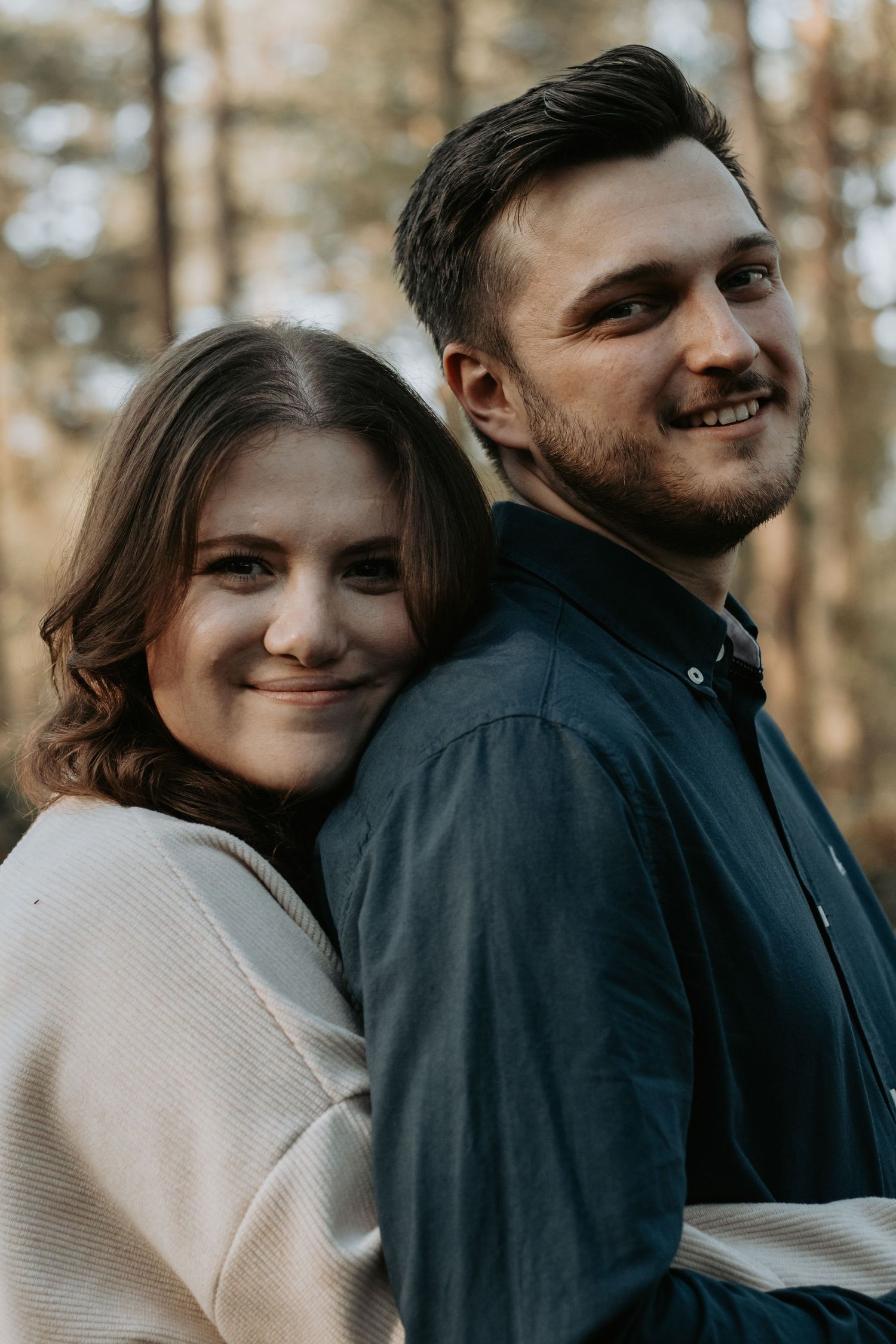 Smiling couple standing back to back in a forest, woman in white sweater and man in dark blue shirt.