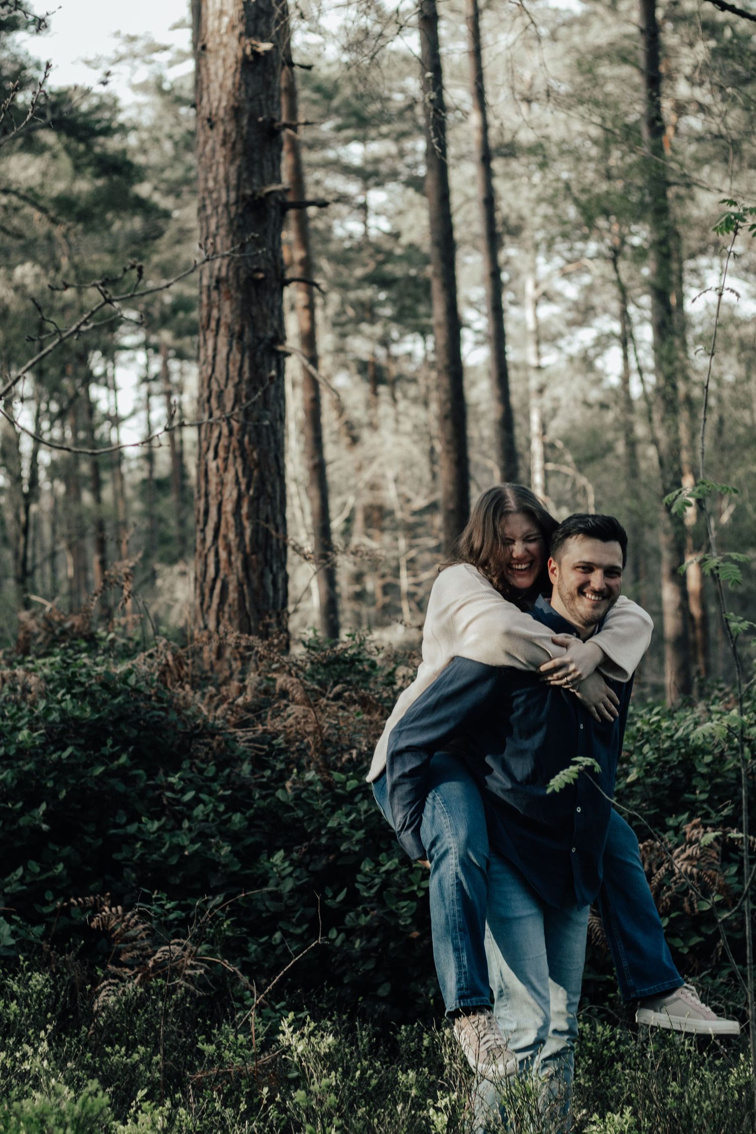 A happy couple shares a piggyback ride in a lush forest, the woman hugging the man from behind as they smile.