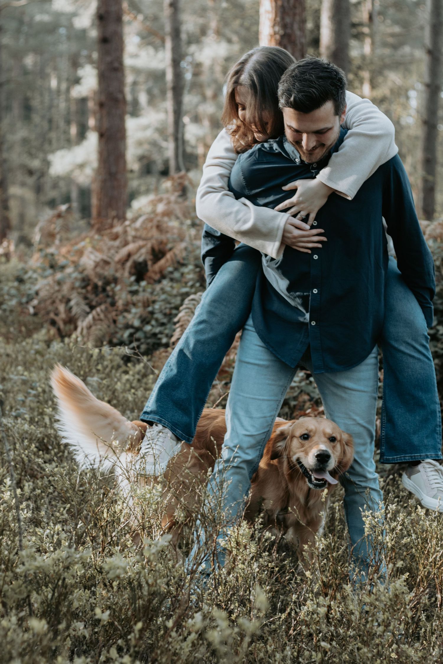 Couple embracing and laughing in a forest with a golden retriever dog among autumn foliage and pine trees.