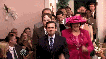 Steve Carell walks with a woman in a pink hat and dress at a festive indoor gathering in The Office TV show.