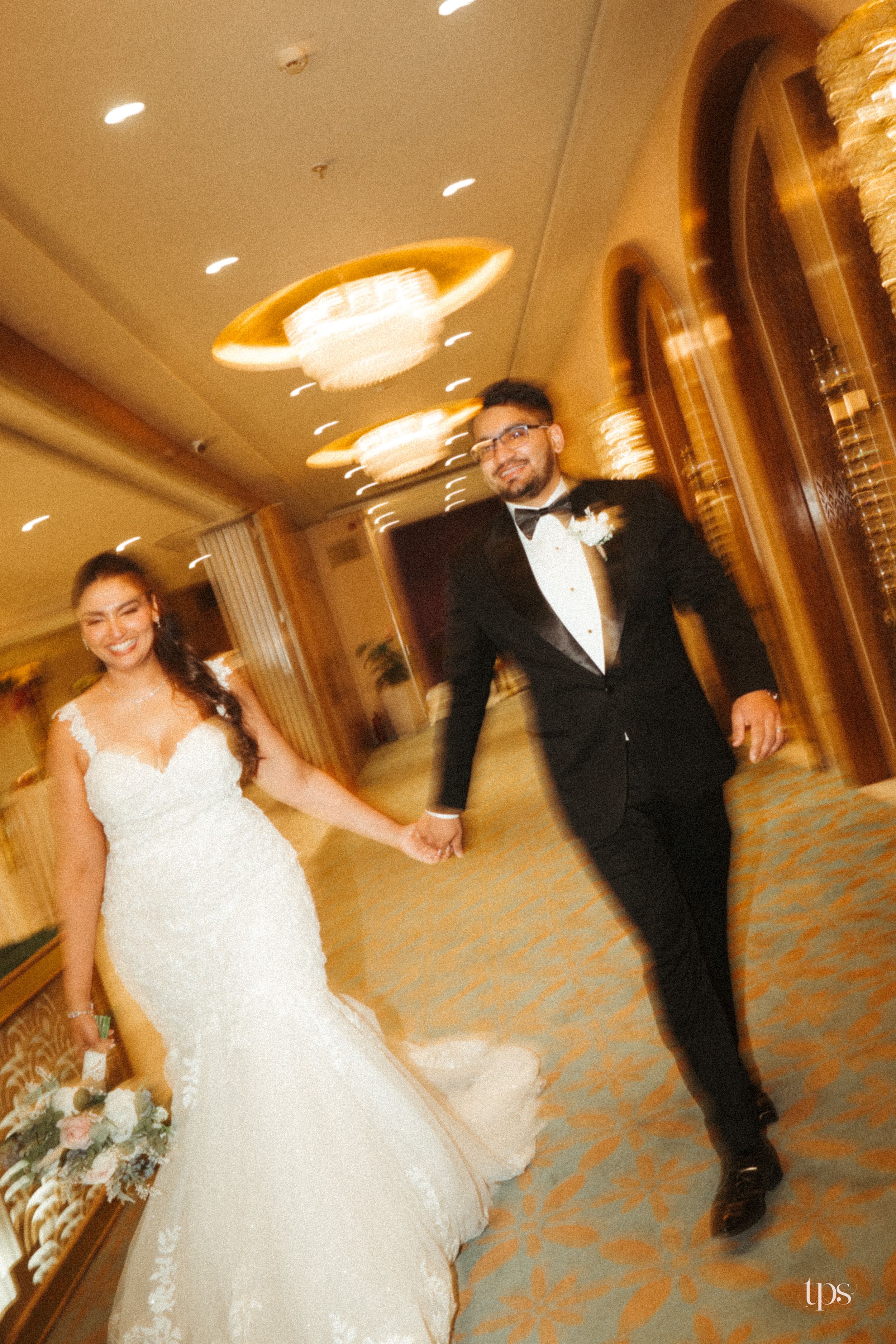 Bride and groom hold hands walking through elegant hotel corridor, bride in lace mermaid dress, groom in black tuxedo.