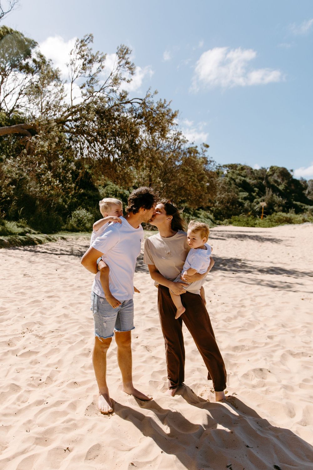 A young family with two toddlers sharing a kiss on a sunny sandy beach with lush green trees behind them.