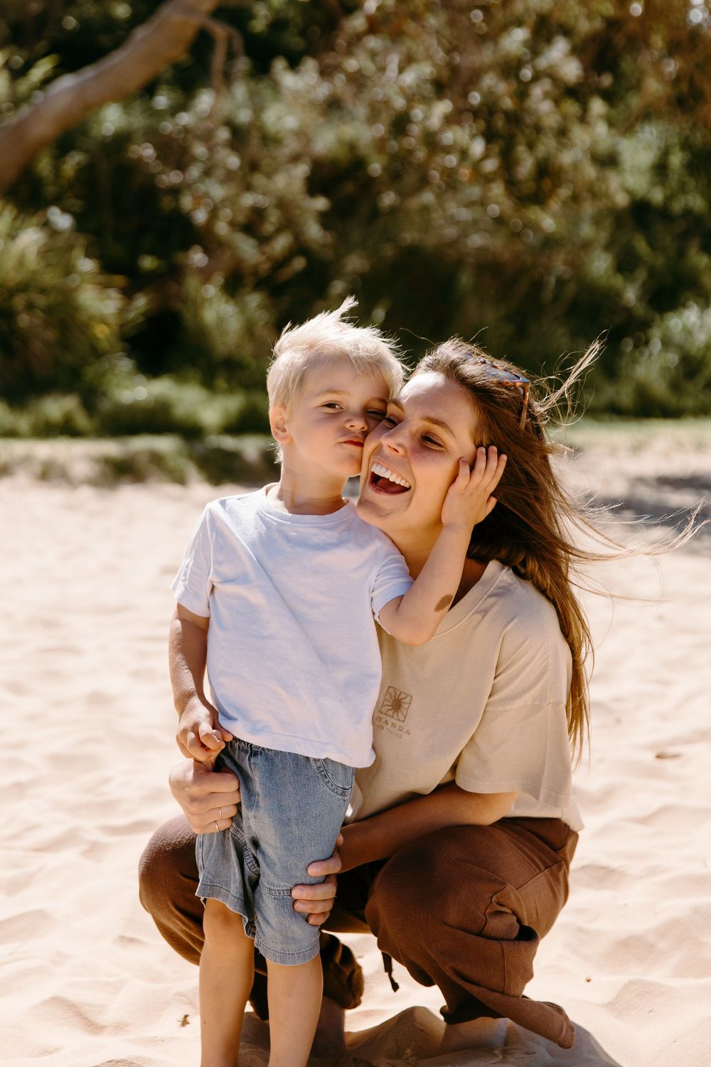 A mother crouches on sandy beach hugging her young blonde son who whispers in her ear.