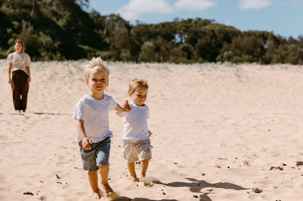 Two young boys in white shirts run on a sunny sandy beach while a woman walks behind them.