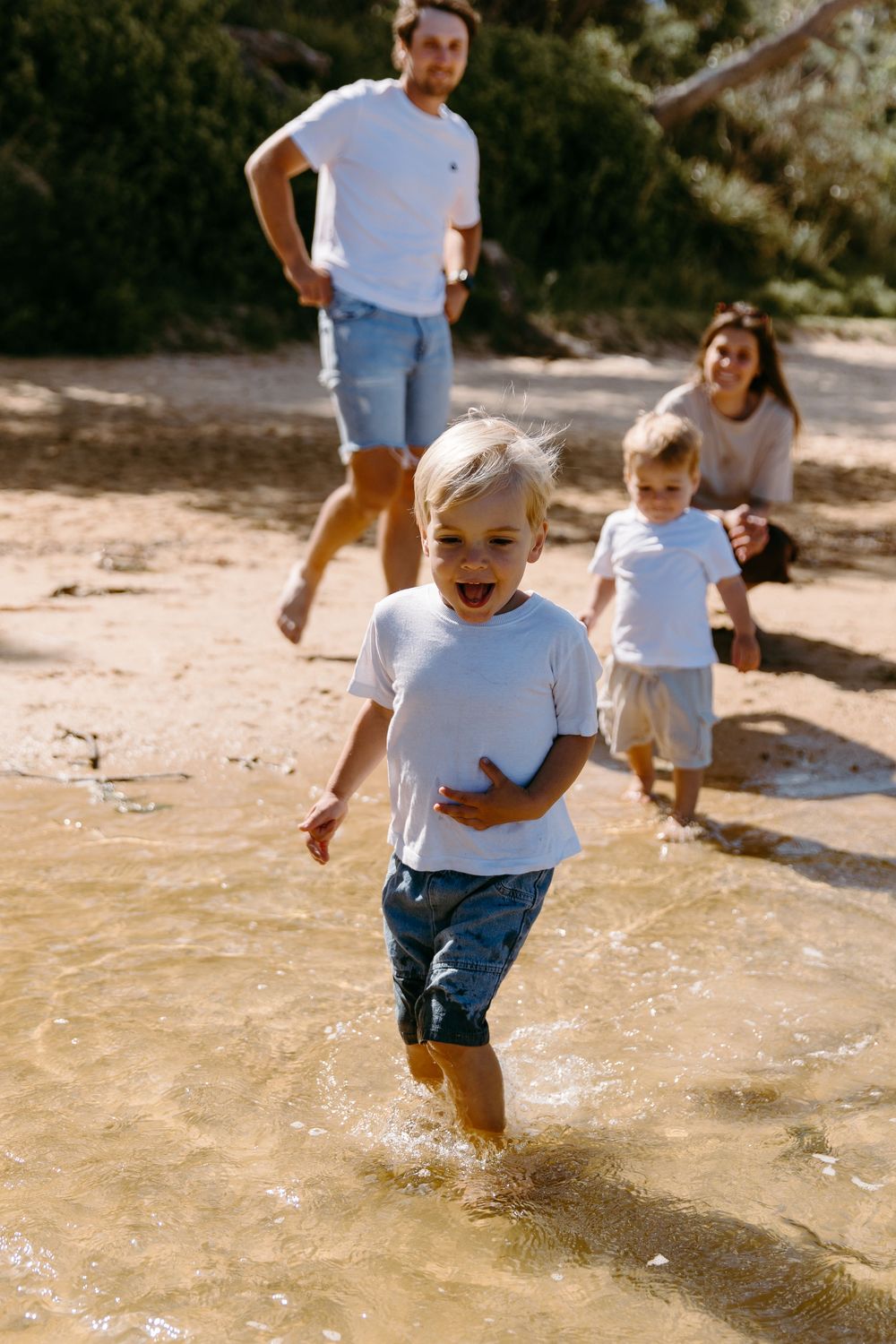 A joyful blonde boy runs through shallow water at a sandy beach while his family watches in the background.