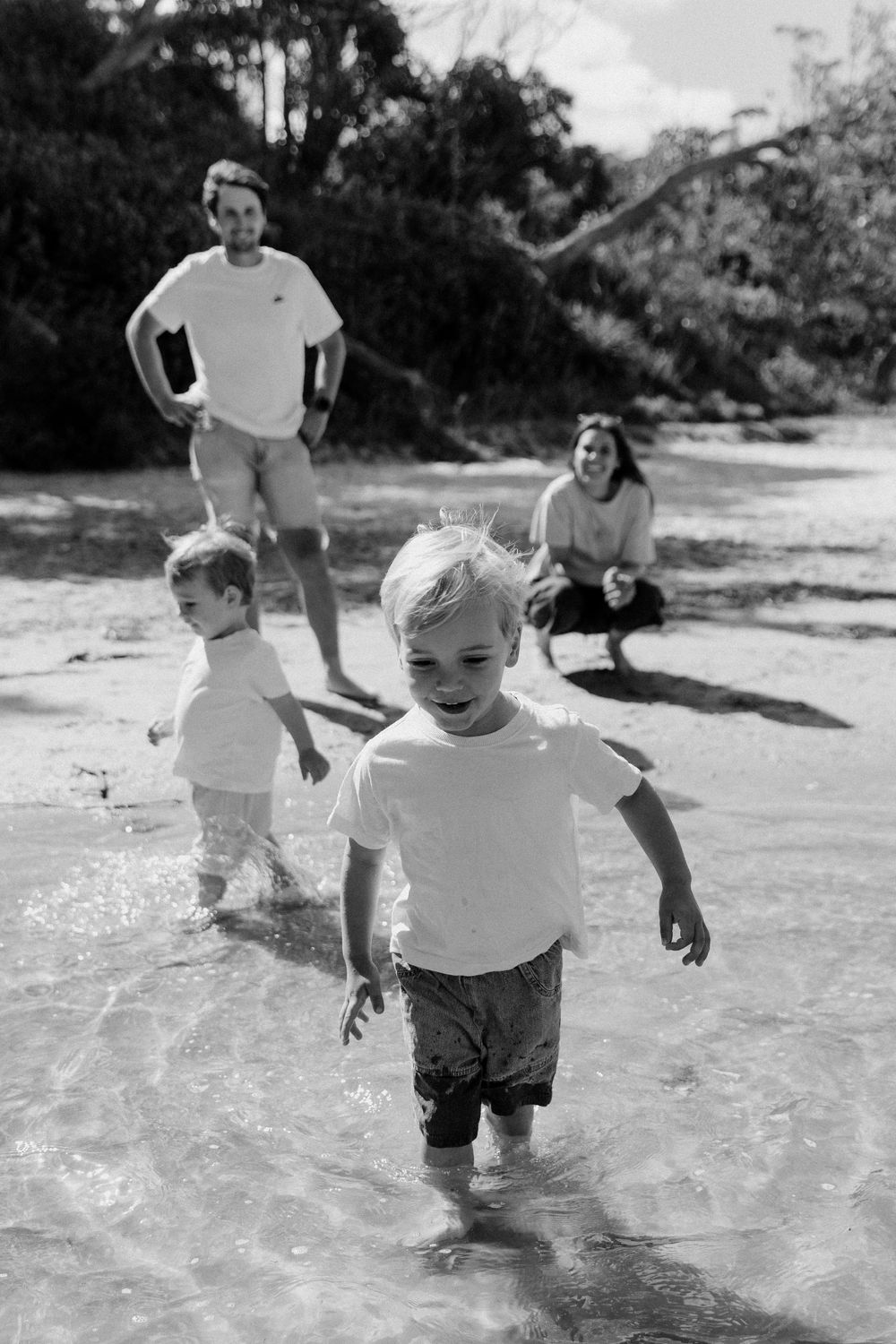 Black and white photo of two young boys wading in shallow water with parents watching.