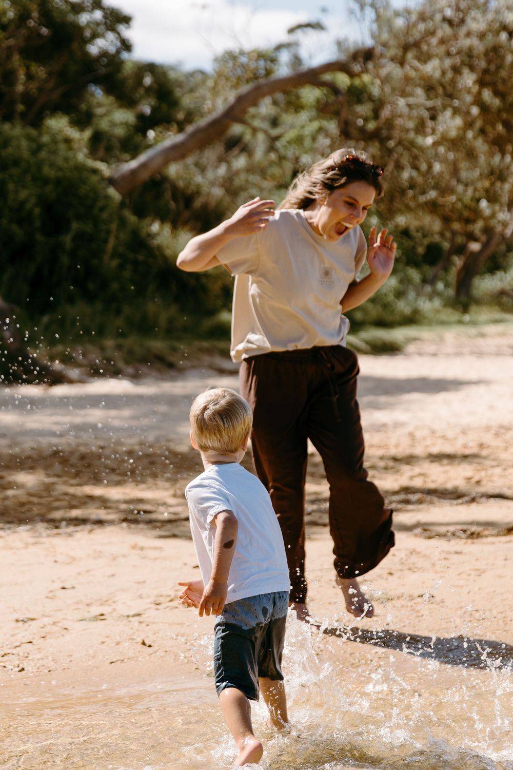 A woman and young child joyfully splashing in shallow water on a sunny beach surrounded by lush green trees.