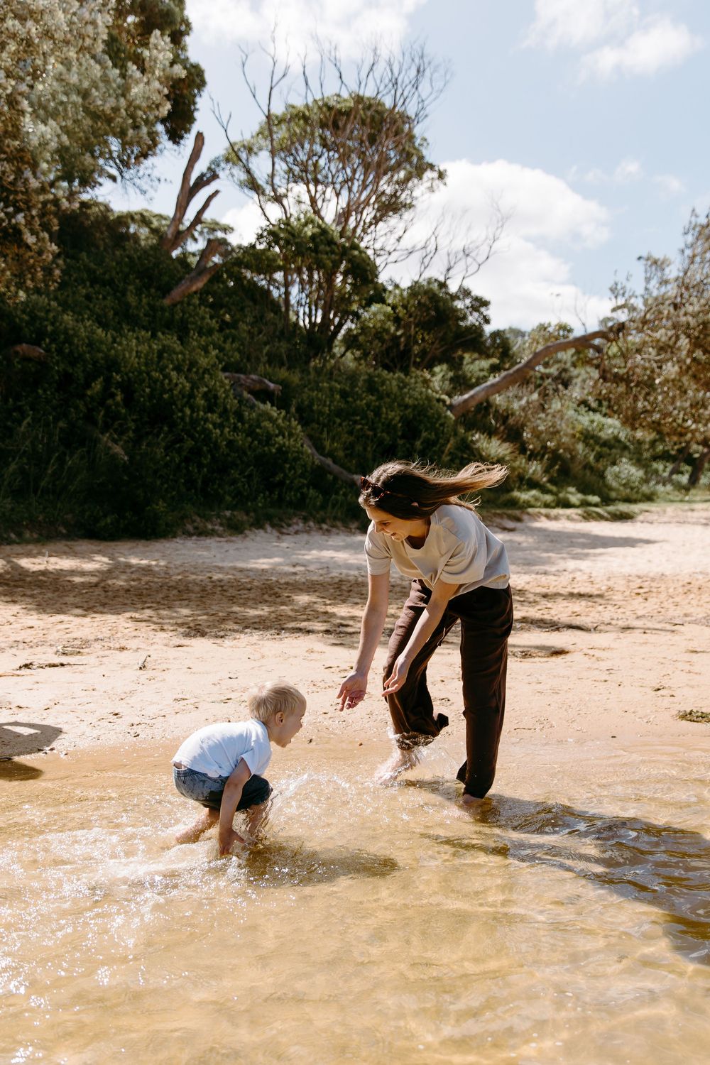 A mother and toddler play together in shallow water on a sandy beach surrounded by lush green trees.