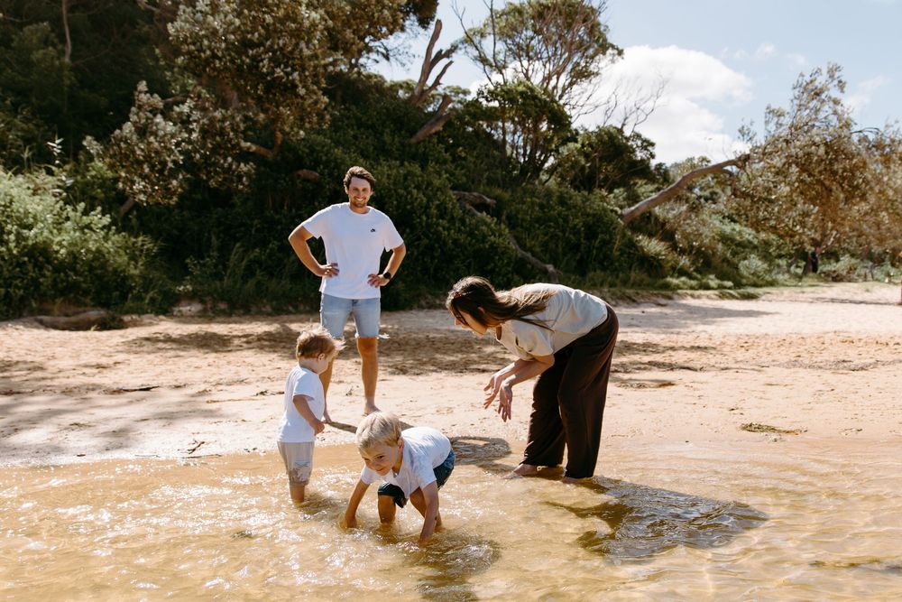 Family playing on a sandy beach shore with children splashing in shallow water surrounded by lush green trees.