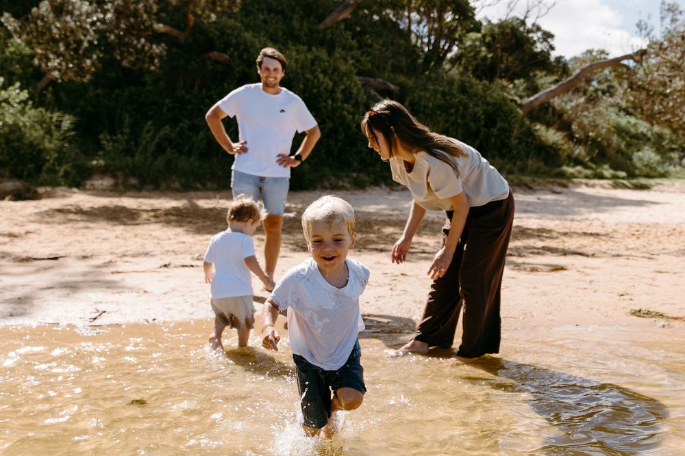Family playing in shallow water on a sandy beach, two young children splashing while parents watch and smile outdoors.