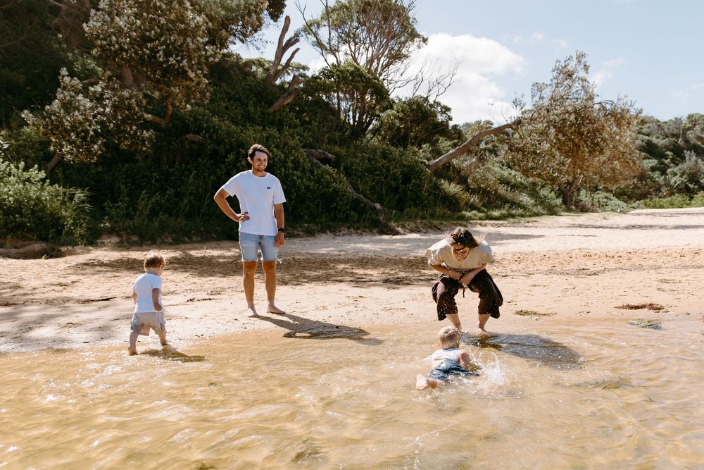 Family playing on a sandy beach near shallow water surrounded by lush green trees on a sunny day.