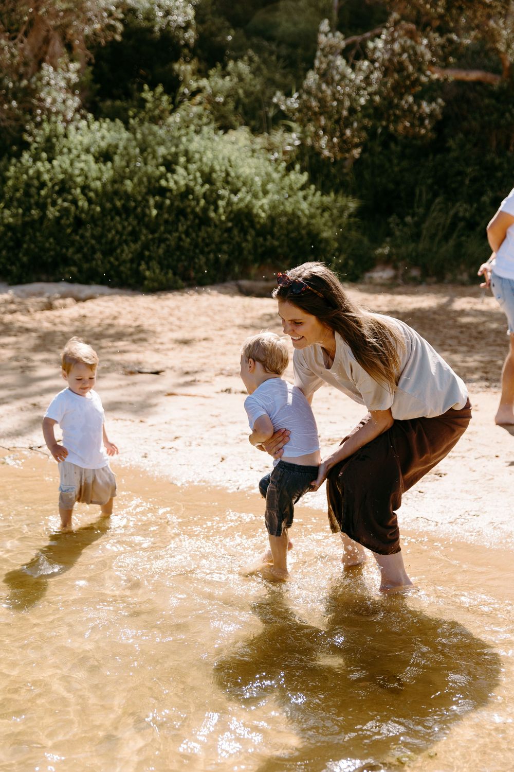 A smiling woman helps two toddlers play in shallow water on a sunny sandy beach surrounded by greenery.