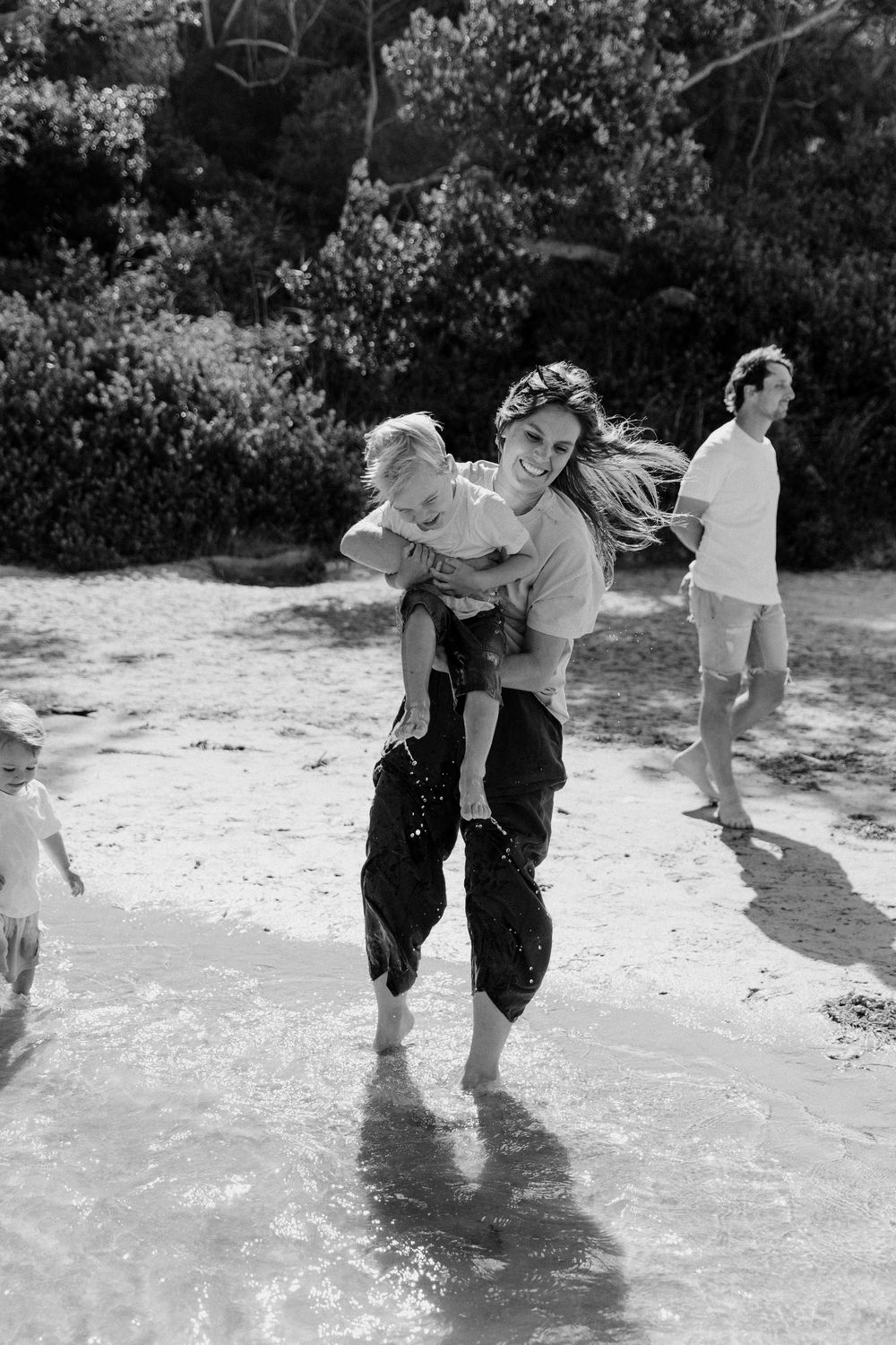 Woman laughing while giving child piggyback ride on beach with family in black and white photo.