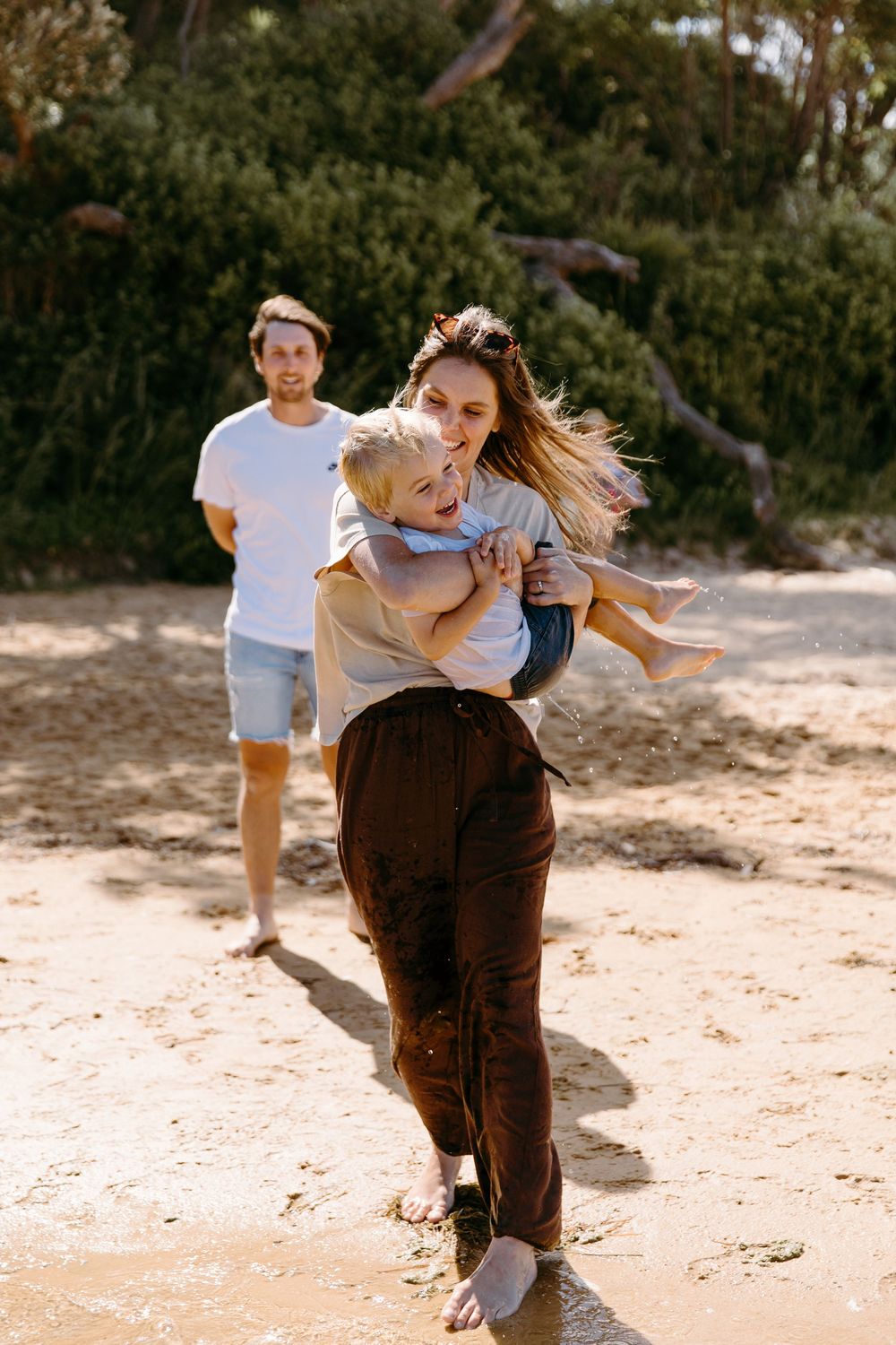 Mother carries laughing child on sandy beach while father walks behind in warm sunlight.