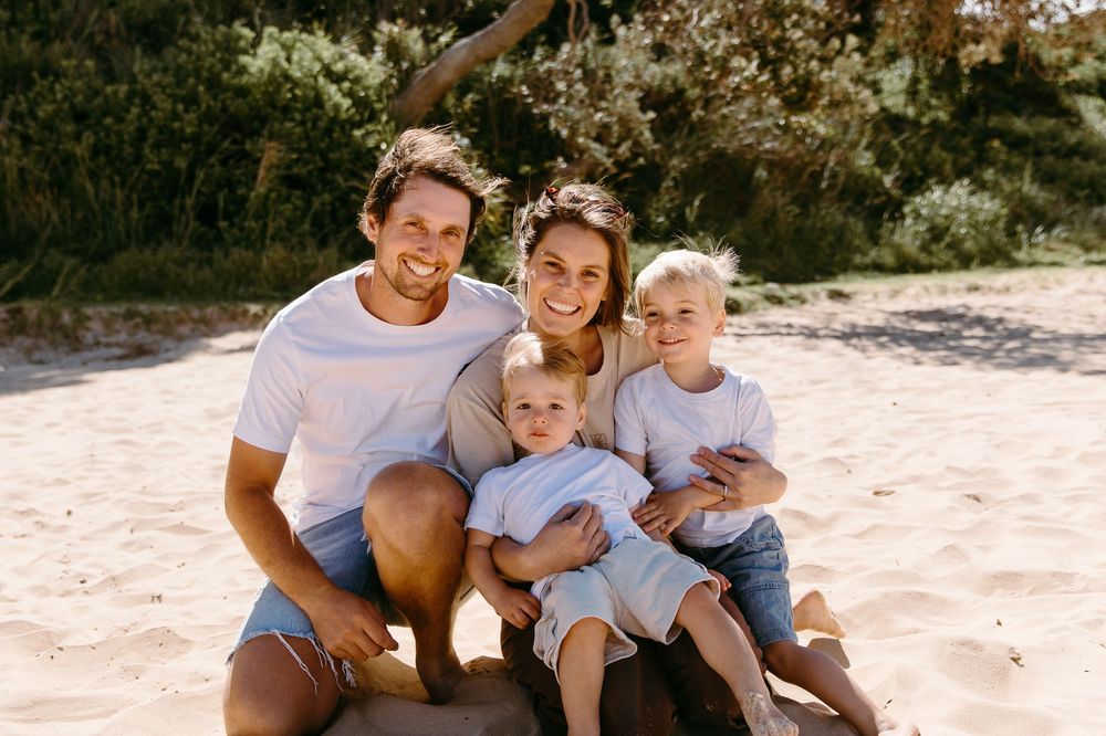 Happy family of four smiling together on a sandy beach, parents with two young blonde children on a sunny day.