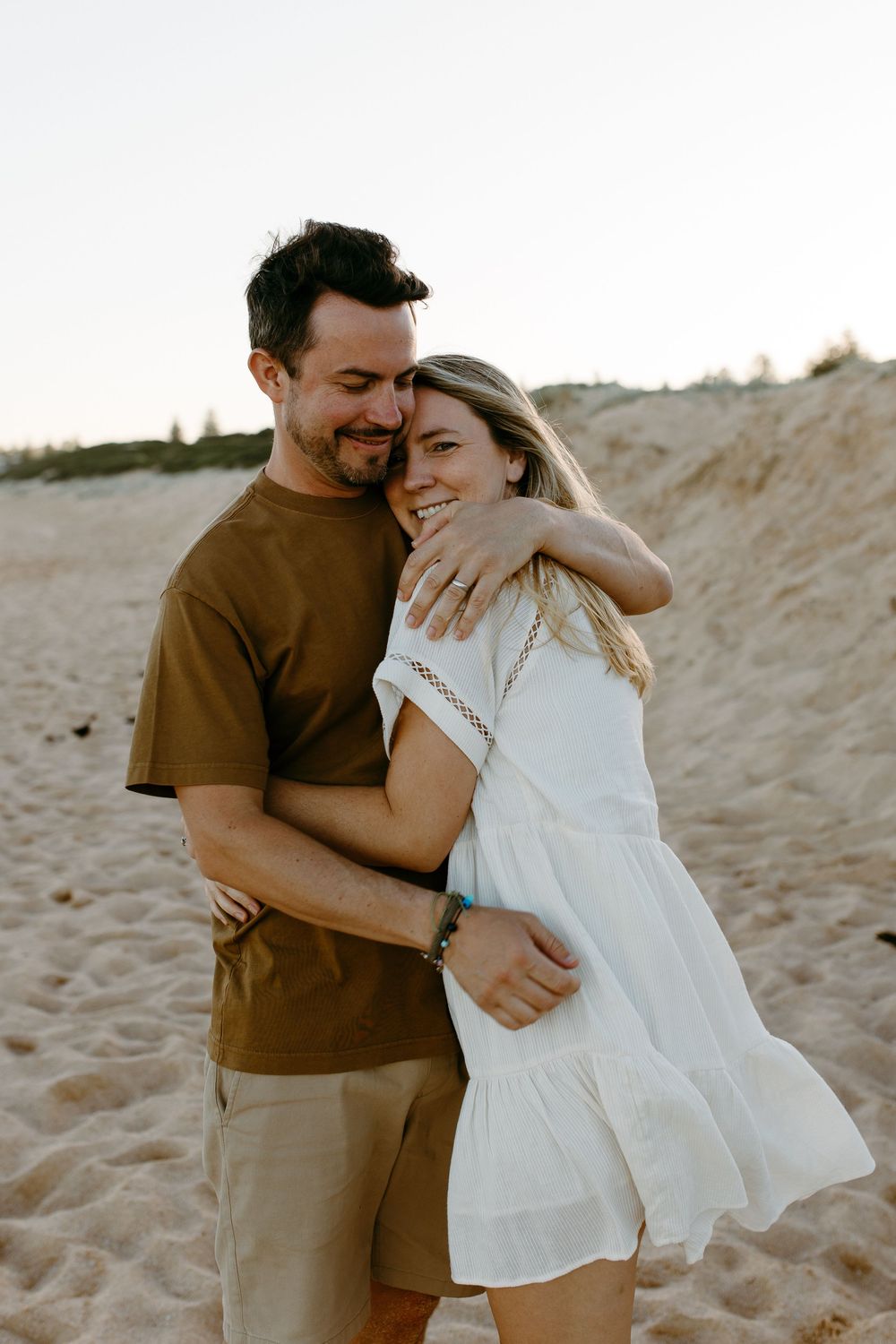 Happy couple embracing on sandy beach, woman in white dress and man in brown shirt, smiling together at sunset.