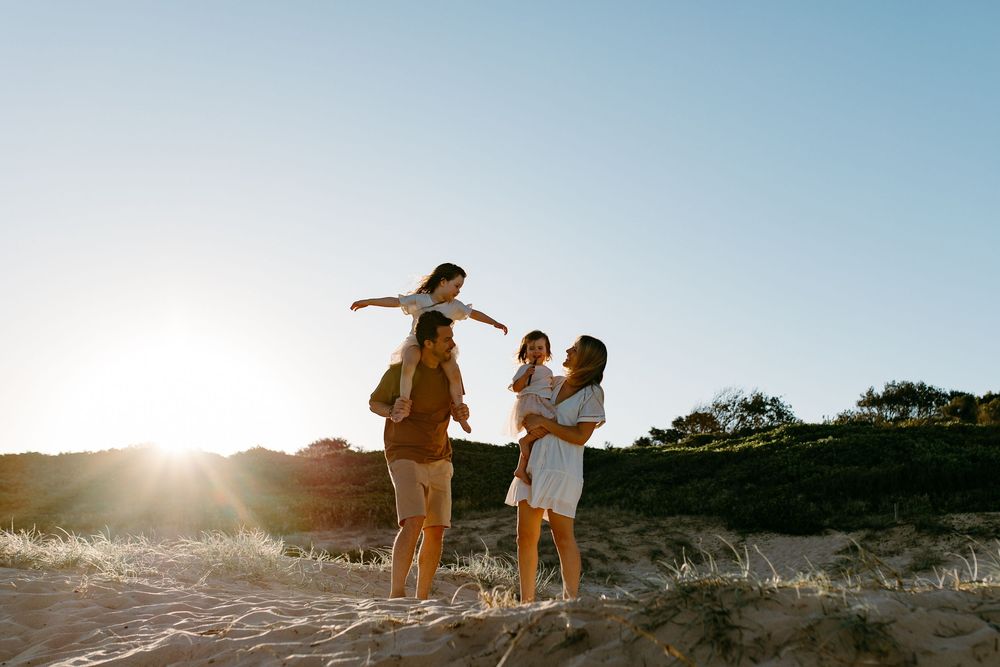 Family enjoying a sunny beach day, with parents lifting children in the air on golden sand dunes at sunset.